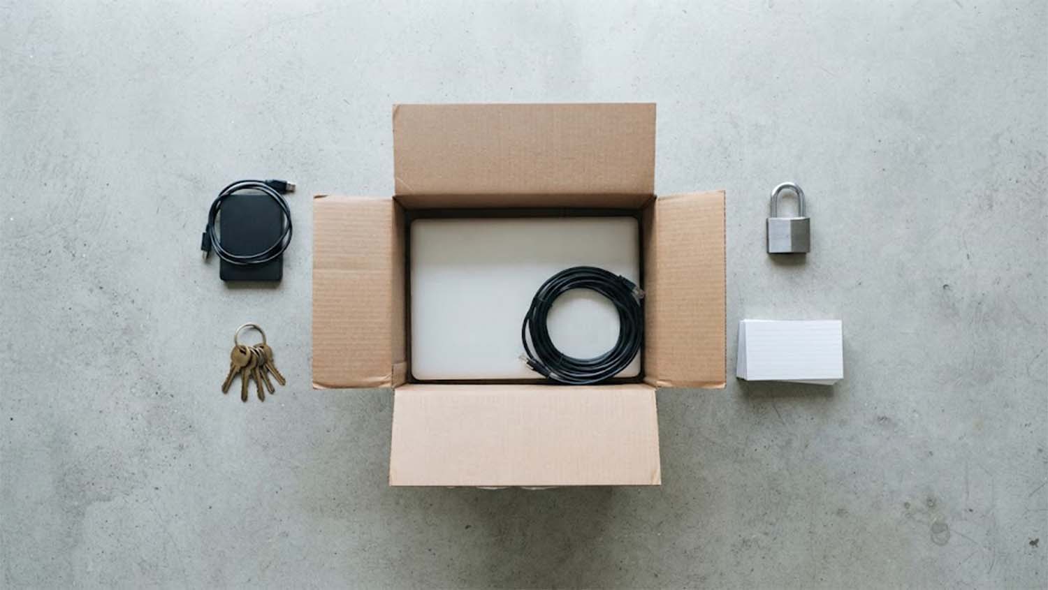 An open cardboard moving box on a grey concrete floor holds a closed laptop and a coiled network cable, while an external hard drive, a ring of keys, a padlock and a stack of index cards sit outside it