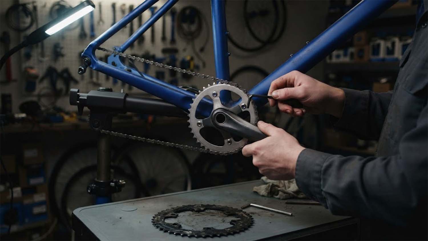 Hands in a dark grey work shirt use a hex key to fit a new chainring to a cobalt blue bicycle frame on a repair stand, with the worn chainring lying on the workbench below