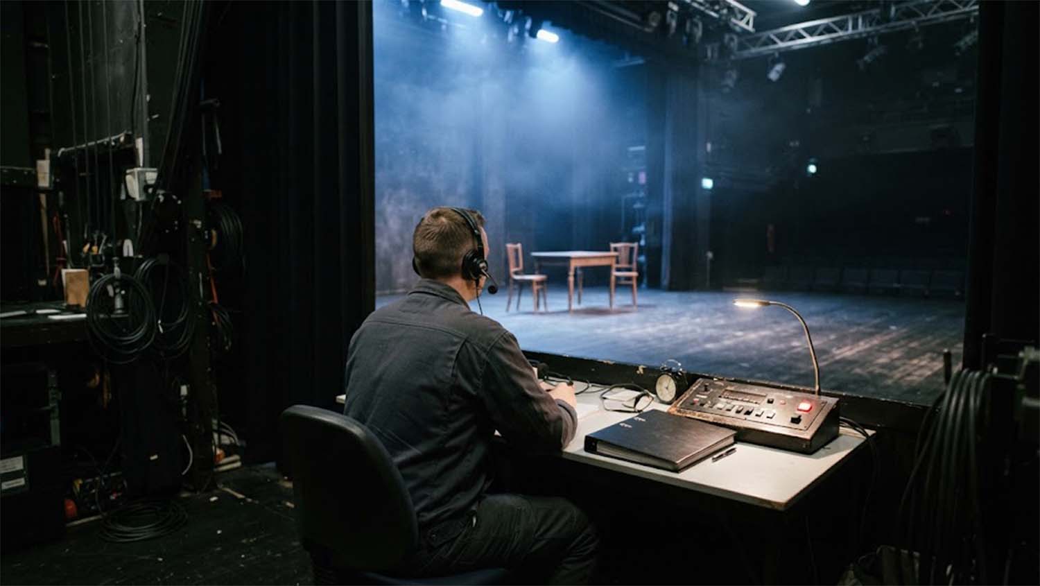 A stage manager wearing a headset sits at a desk in the wings of a small theater, with a black binder, a desk clock, a gooseneck lamp and a cue control panel, looking out at an empty stage lit in hazy blue light with a wooden table and two chairs