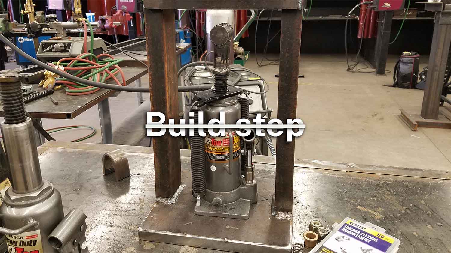 A hand-welded hydraulic press in a metal fabrication shop, with the words 'Build step' crossed out overlaid on the image