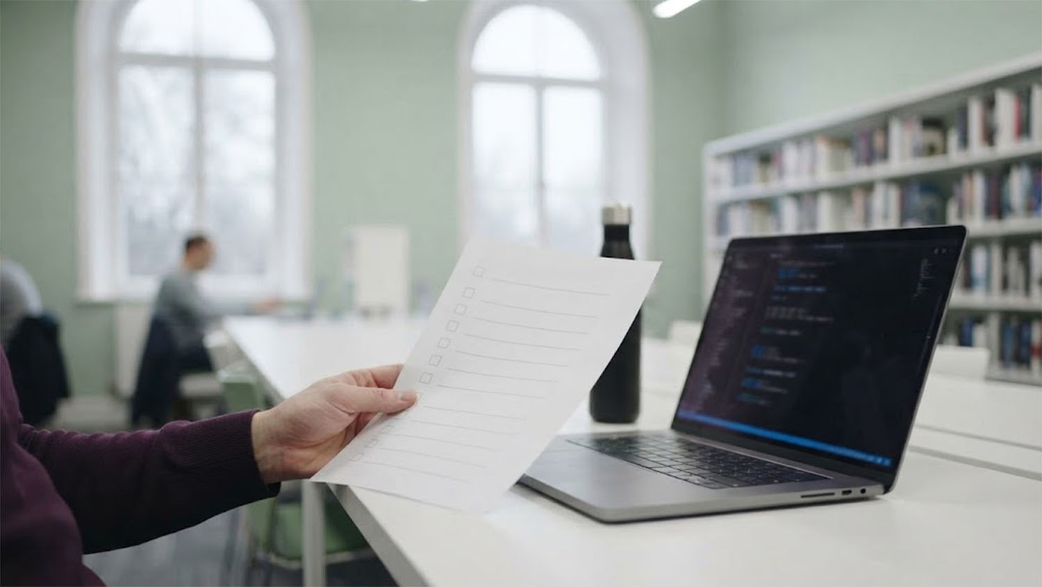A hand in a plum sleeve holds a printed checklist of empty checkboxes beside an open laptop showing a code editor, on a white table in a library reading room