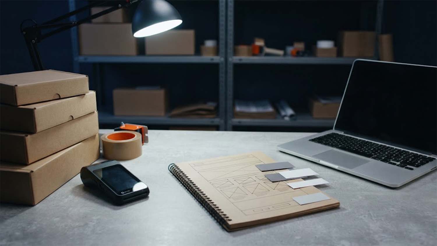 A packing counter at night with a stack of kraft mailer boxes, a roll of packing tape, a card payment terminal, an open laptop and a spiral sketchbook showing a hand-drawn web page wireframe with grey paper rectangles laid on it, lit by a black desk lamp in front of shelves of boxes