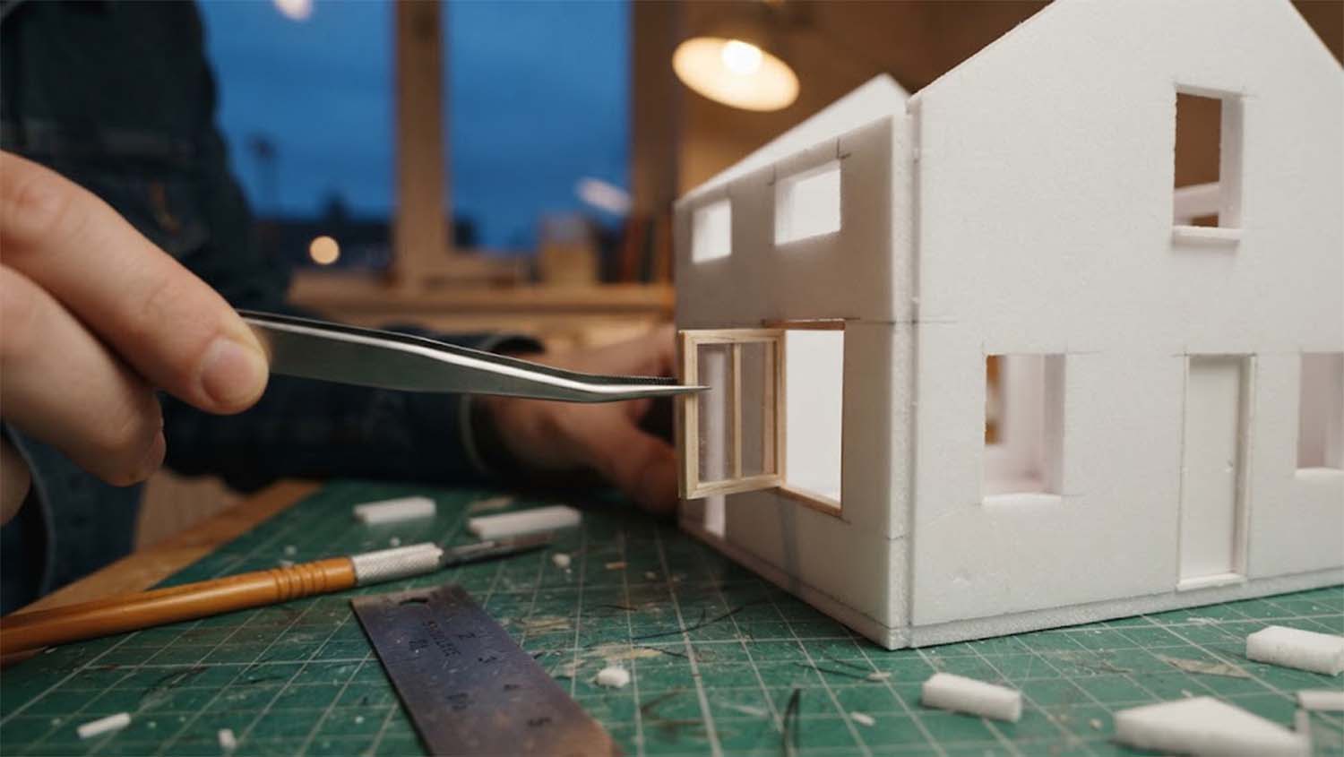 Low close-up of a hand holding fine steel tweezers that place a small wooden window frame into an opening in a white foam-board model house on a green cutting mat, with a craft knife, a metal ruler and foam offcuts nearby, a warm pendant lamp and a dusk-blue window behind